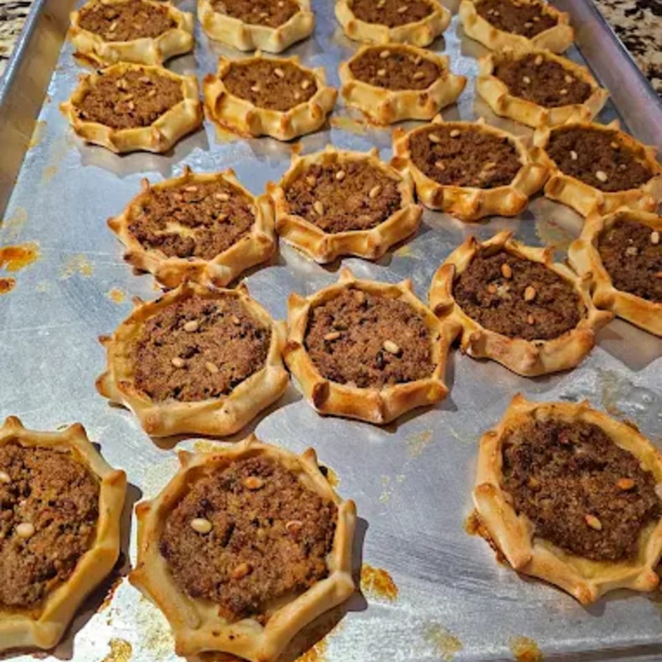 A baking sheet filled with numerous savory meat pies, each topped with pine nuts.