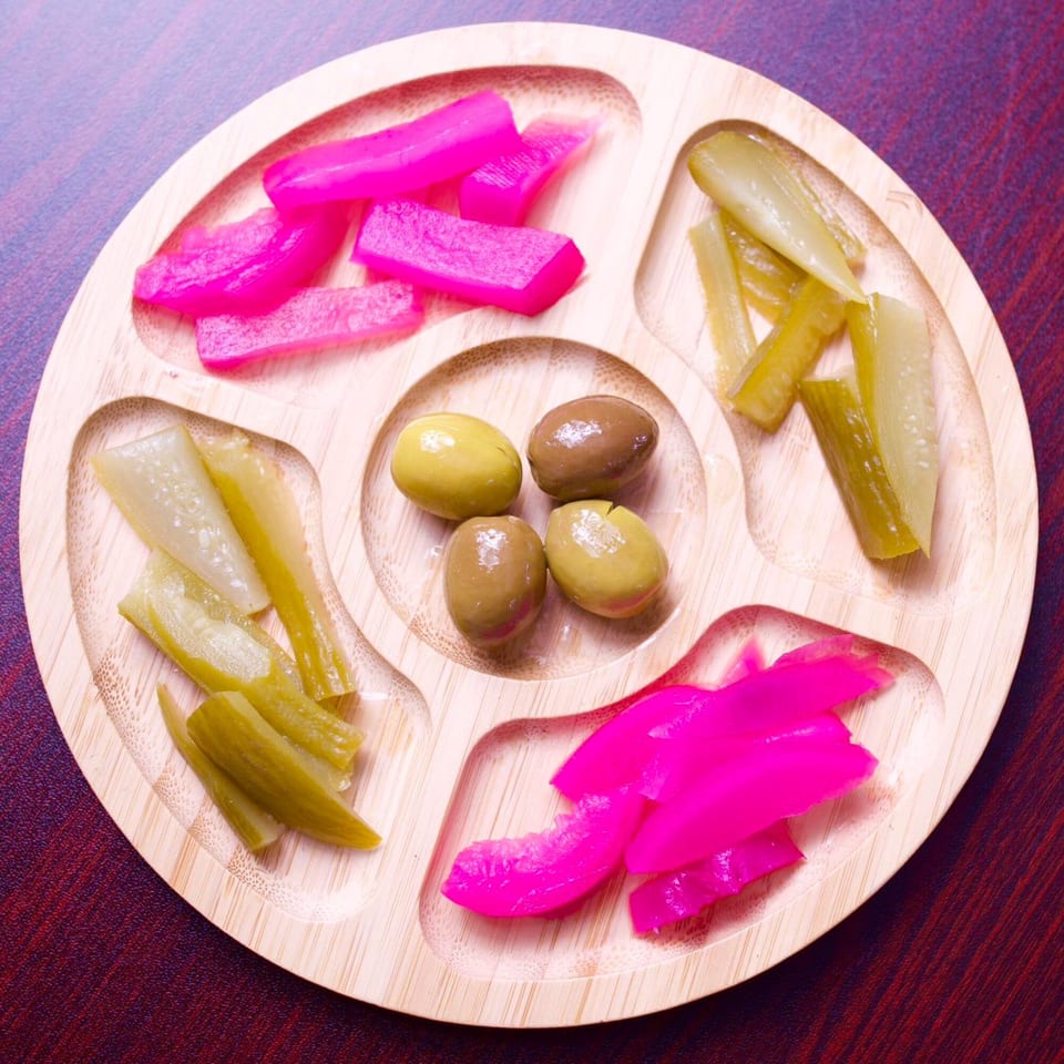 A wooden serving tray with compartments of bright pink pickled turnips, green pickled cucumbers, and olives.