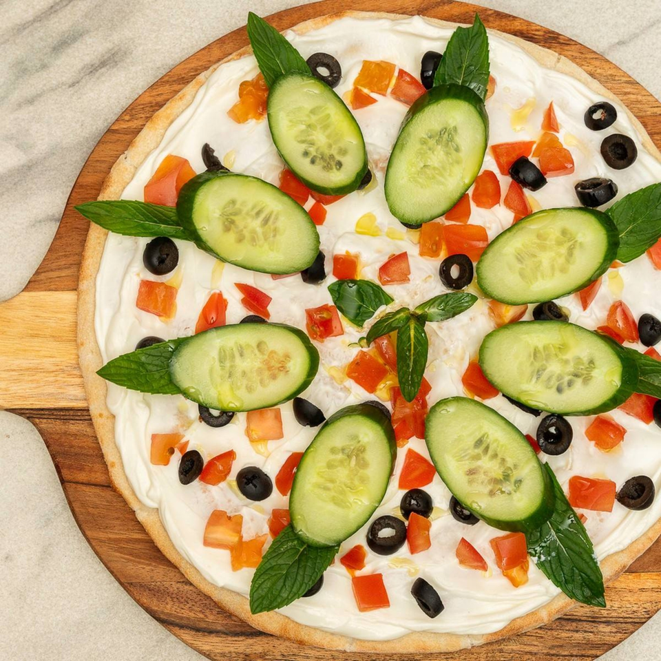 Flatbread topped with white cream, cucumber slices, diced tomatoes, black olives, and mint leaves.